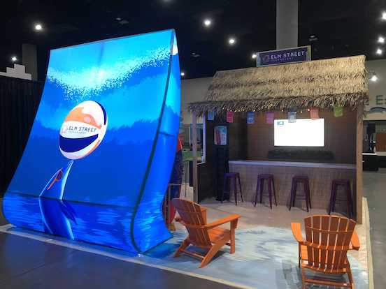 A beach themed trade show booth for Elm Street Technology made by Cardinal Expo. It features an LED wall with ocean waves, marine life, and a branded Elm Street beach ball. The booth is also styled as a tiki hut.