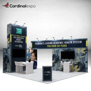 Front-left perspective of a 20x20 trade show booth rental with a tall graphic tower, overhead header beam, TV monitors, high-top meeting tables, and reception desk.